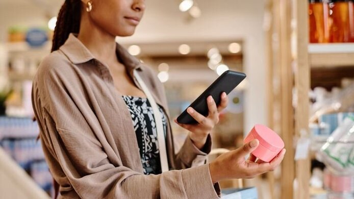 Foto einer Frau im Geschäft, in der rechten Hand ein kosmetisches Produkt, in der linken ein Handy